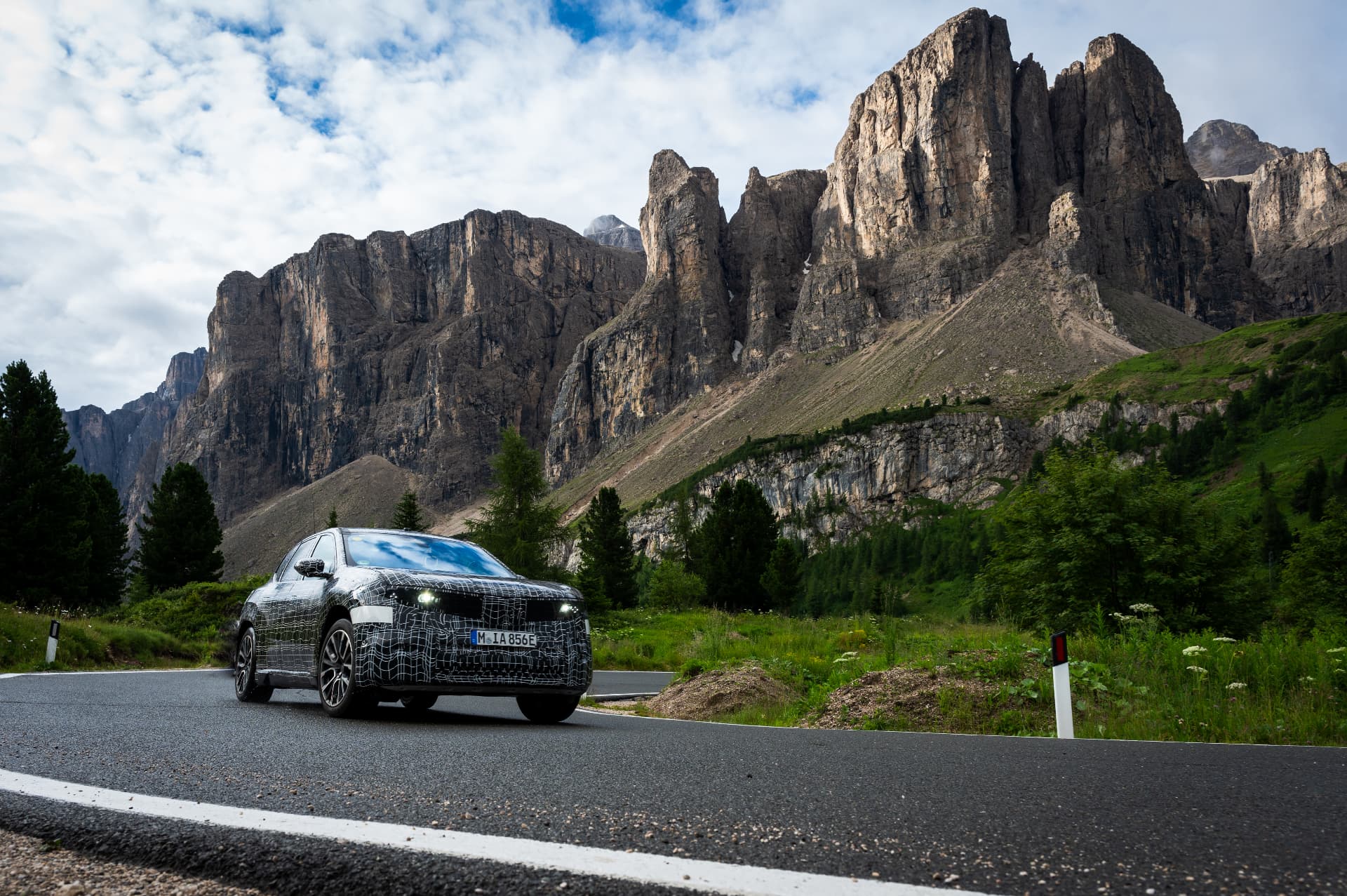 Camouflaged BMW iX3 prototype on a mountain road in front of a steep rock face during testing