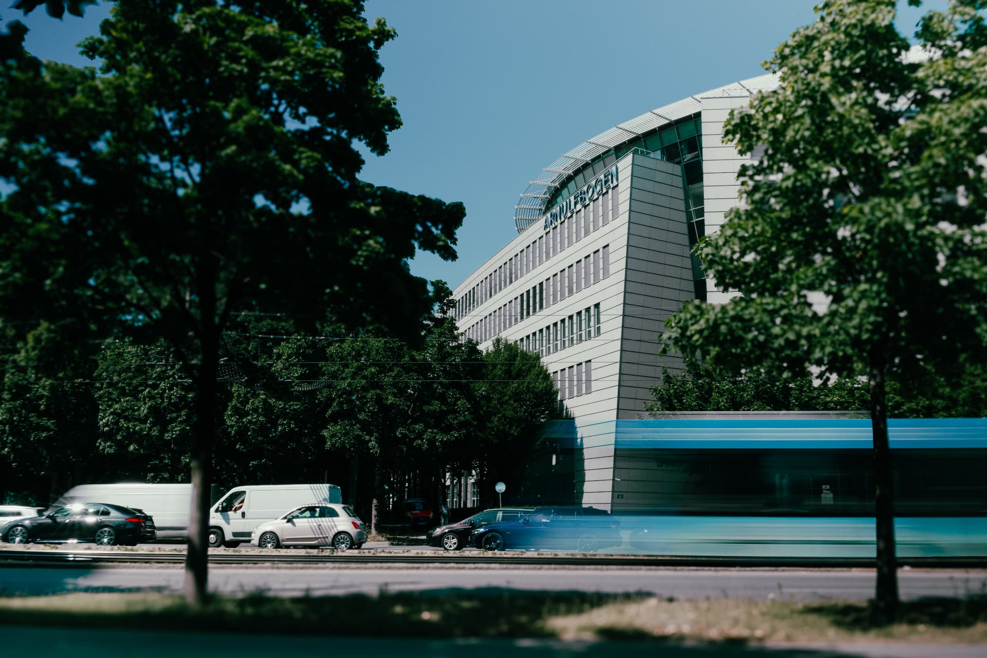 Arnulfbogen seen from across the street through trees, with a blurred blue tram and traffic passing in front of the building