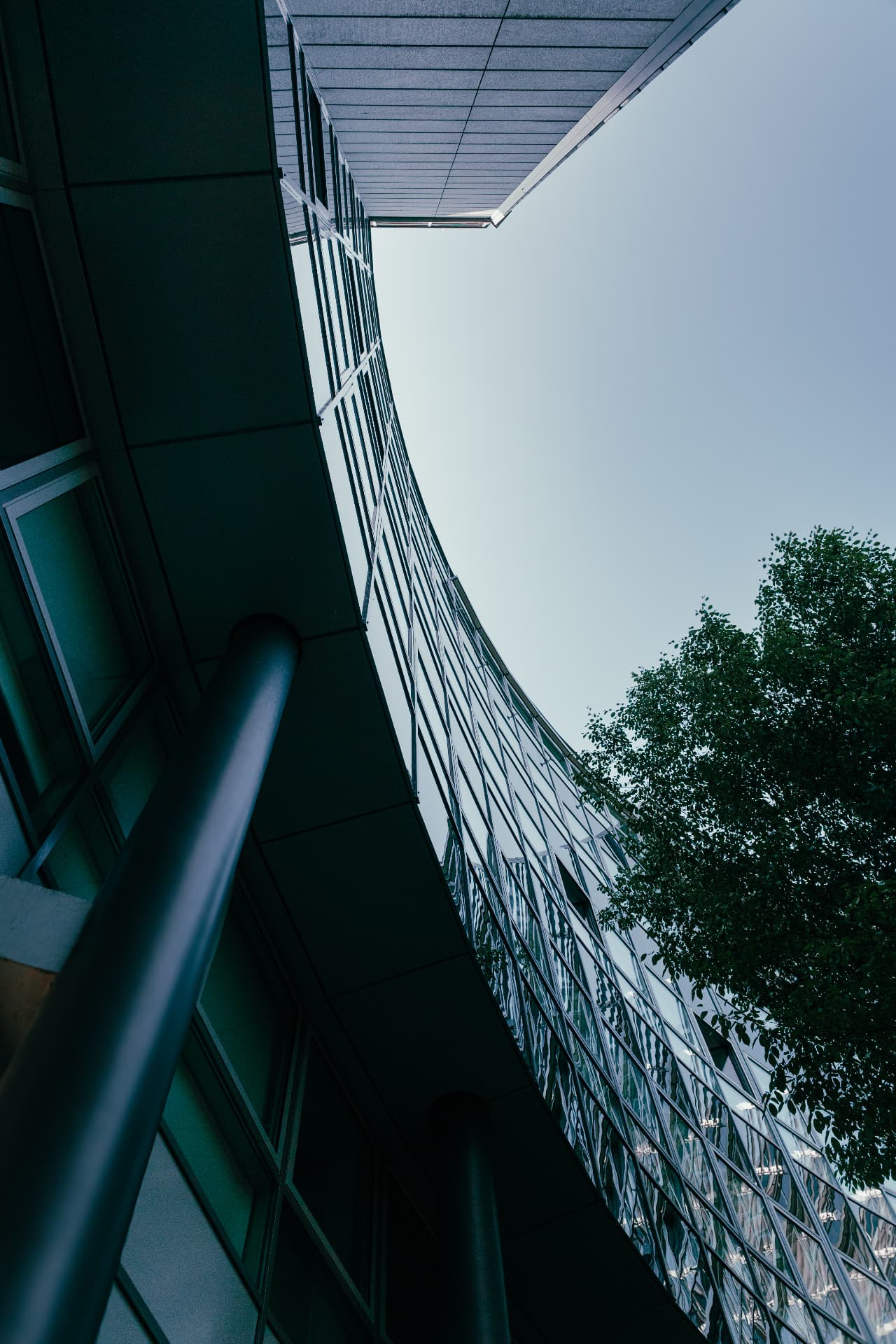 Looking straight up along the curved glass façade of Arnulfbogen, with a steel column and a tree against a pale sky