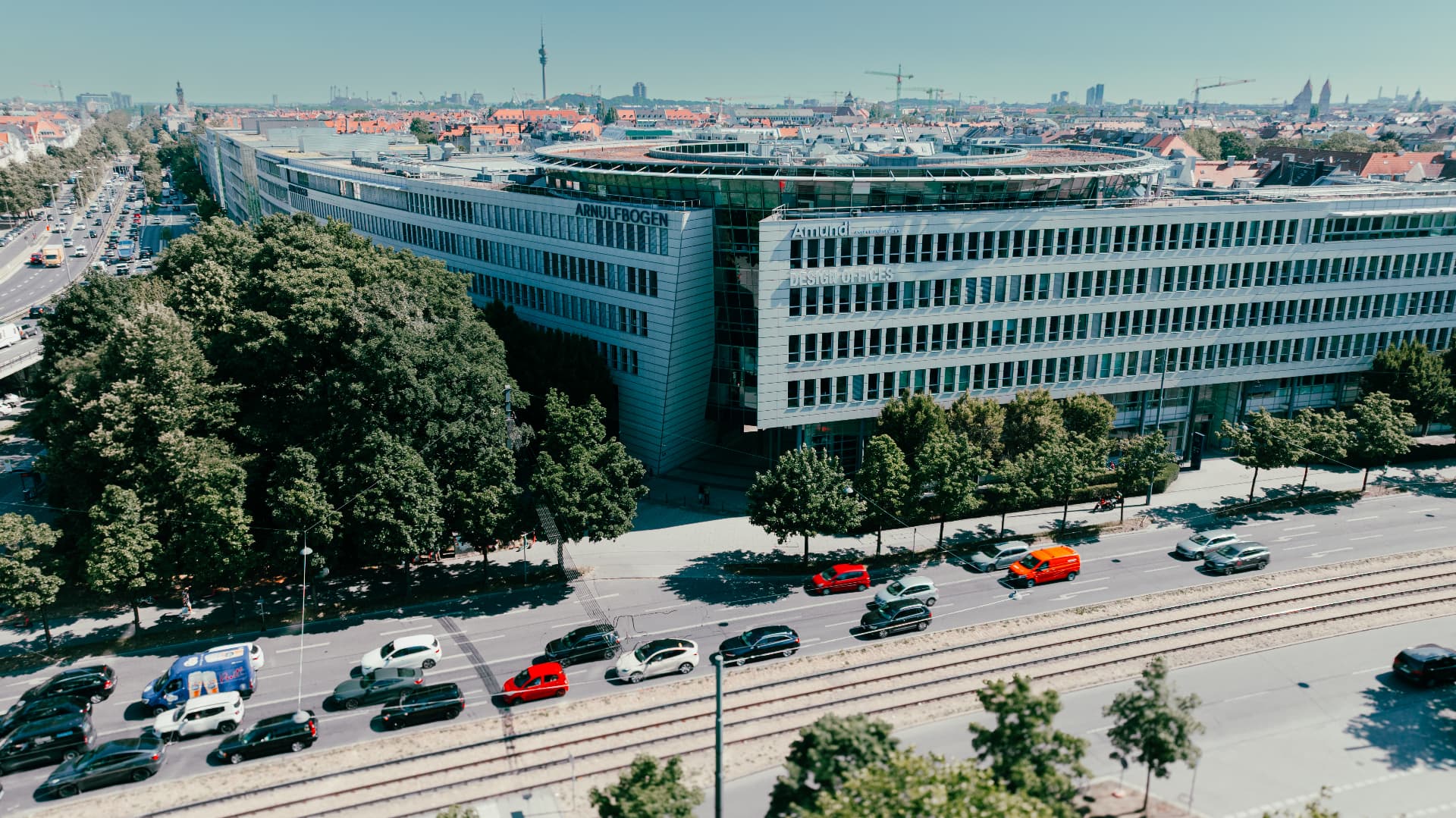 Drone view of the Arnulfbogen building along Arnulfstraße, with traffic, tram tracks and the Munich skyline and Olympic Tower beyond