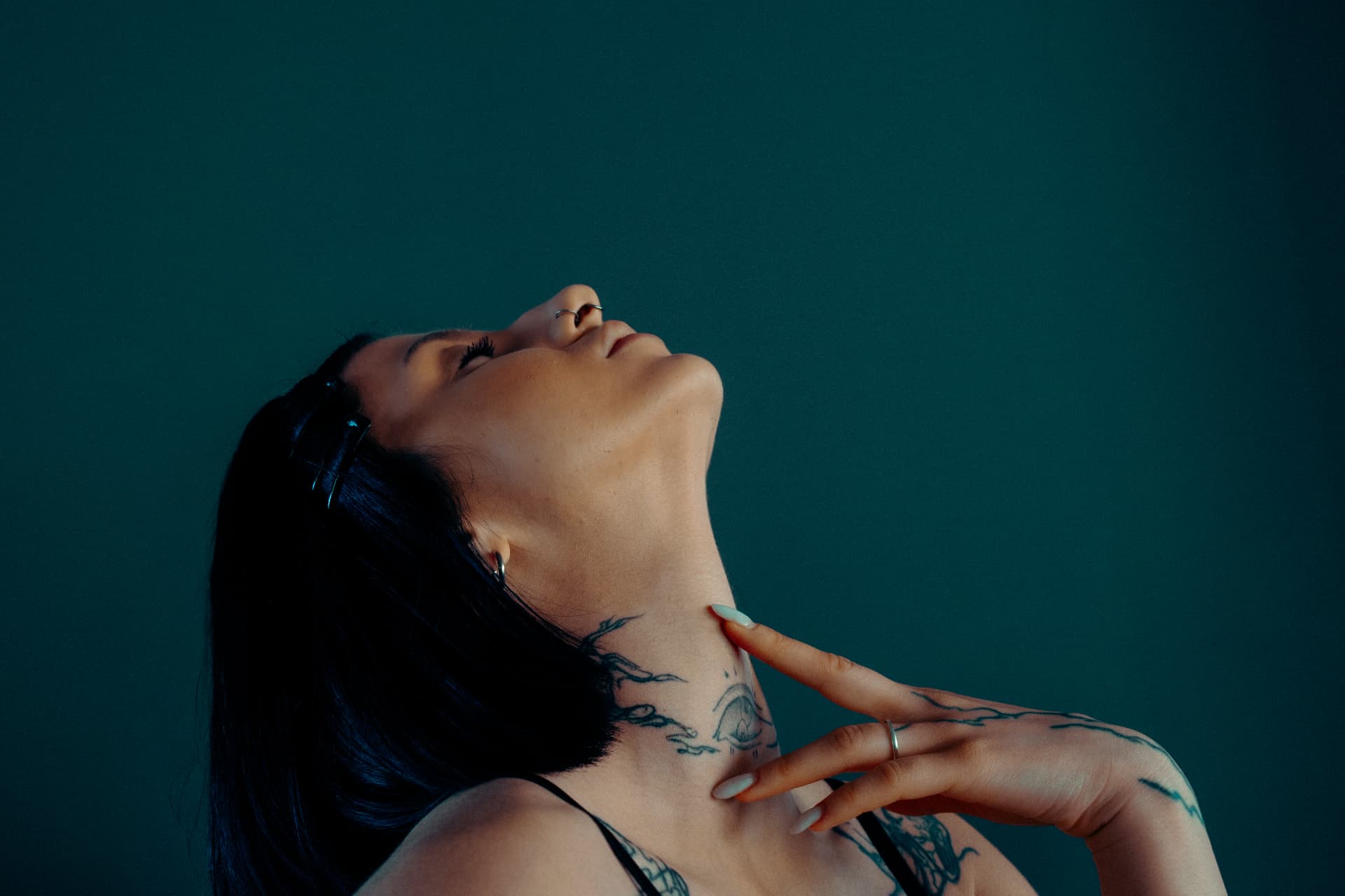 Close-up profile of a model with neck tattoos tilting her head back against a dark green wall