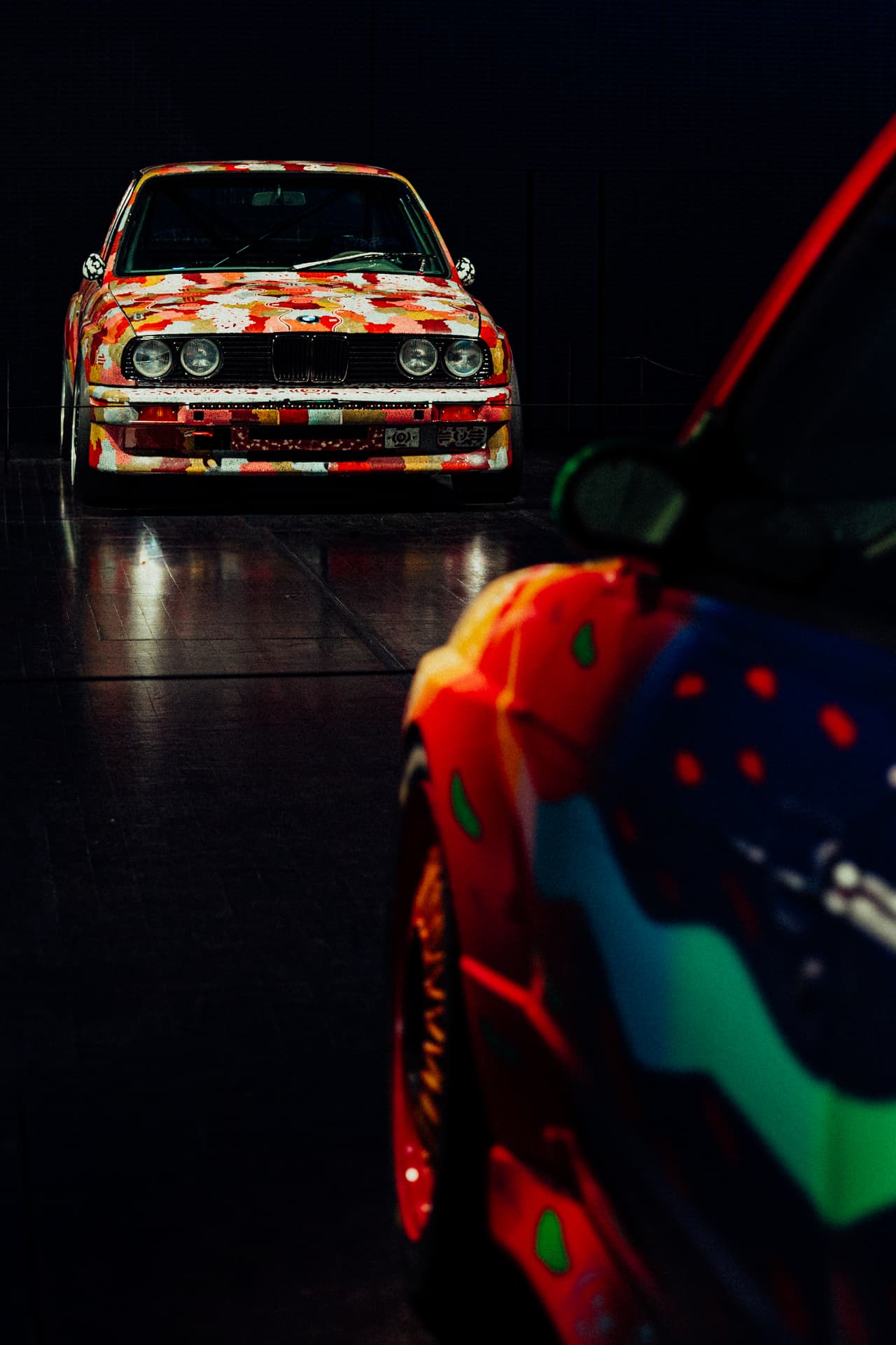 Portrait composition of two vividly painted BMW Art Cars in a dark exhibition space