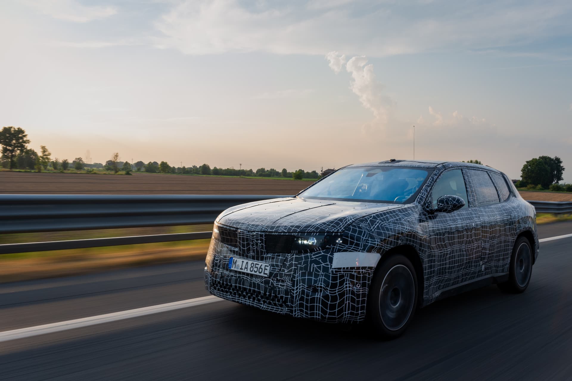 Camouflaged BMW iX3 prototype driving on the highway at sunset during testing