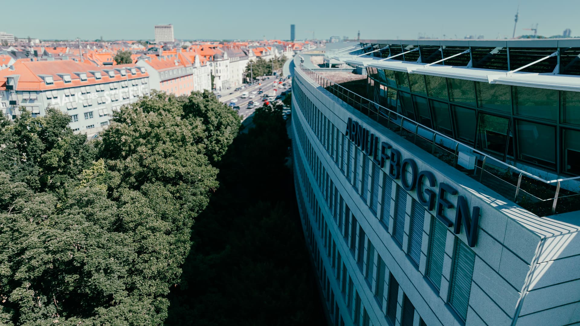 Drone view along the curving Arnulfbogen façade and its lettering, with a line of trees and the Munich rooftops stretching into the distance