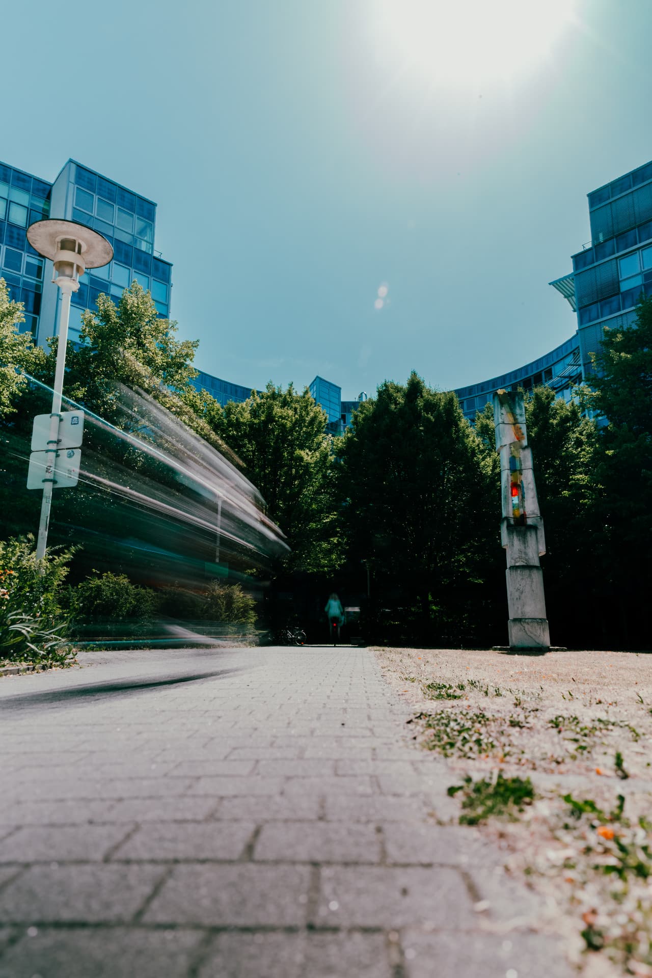 Low-angle view along a paved path into the Arnulfbogen courtyard, with a blurred tram passing and a sculptural column beneath the curved glass façades