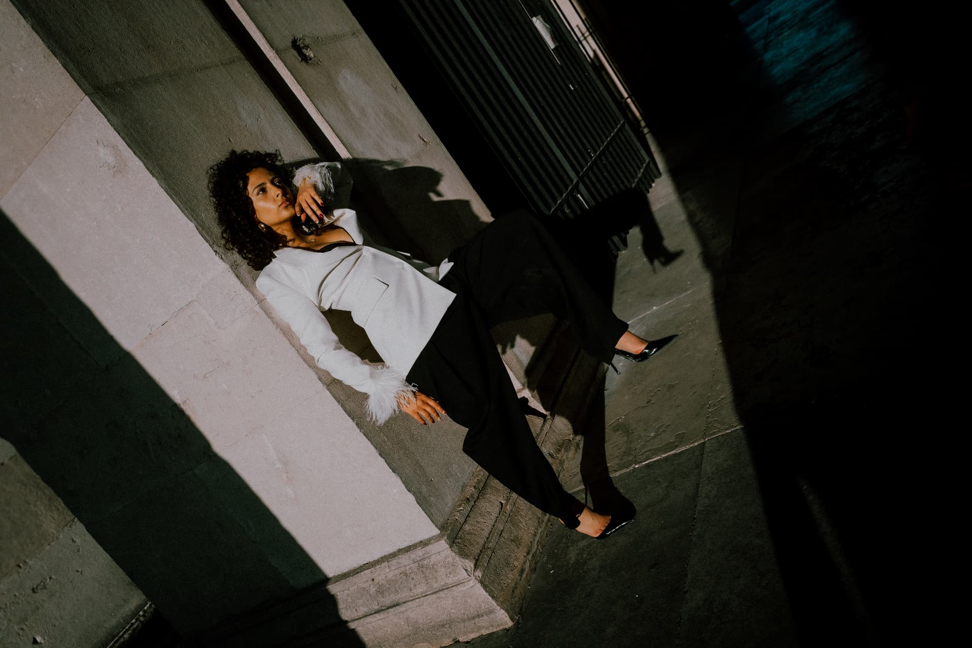 Model in a white blazer with feather cuffs reclining against a stone wall, shot from a high angle