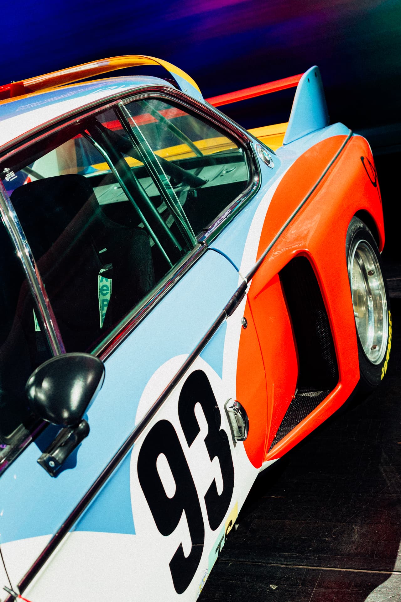 Rear view of a blue and orange BMW Art Car number 93 with rear wing in a dark exhibition space