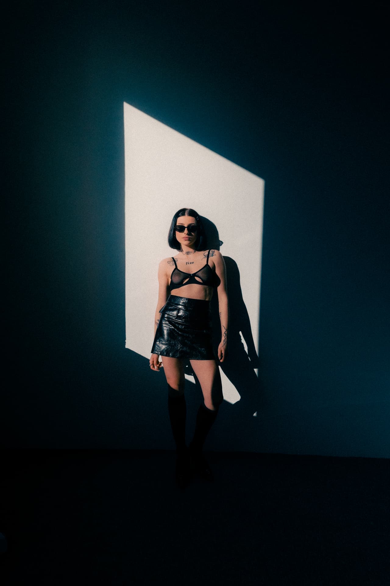 Model in a leather skirt standing inside a beam of light against a dark wall