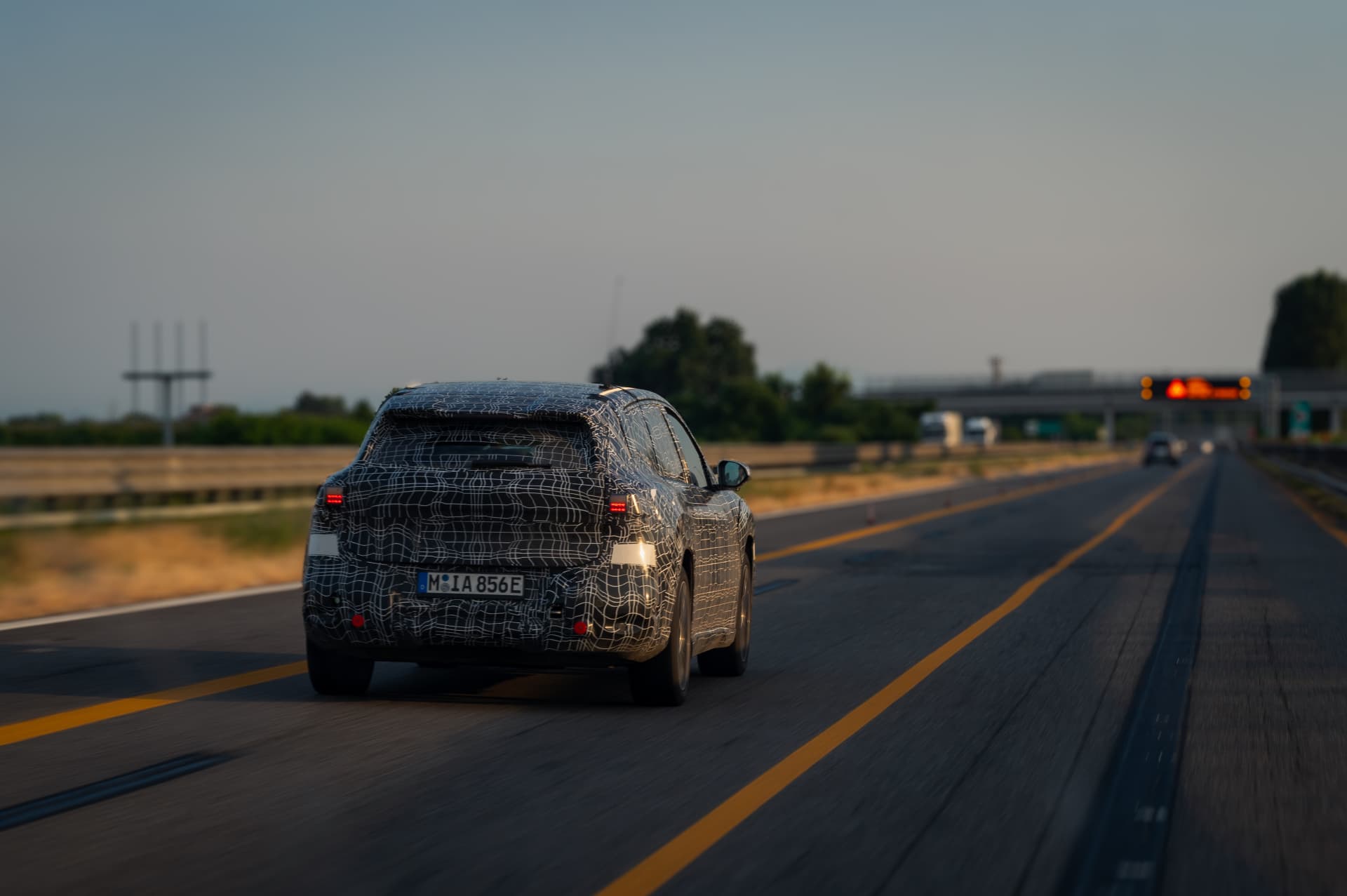 Rear view of a camouflaged BMW iX3 prototype on the highway at dusk during testing