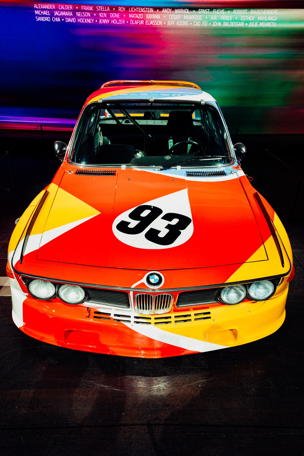 Front view of the orange, yellow and red BMW Art Car number 93 beneath a wall of artist names at BMW Welt