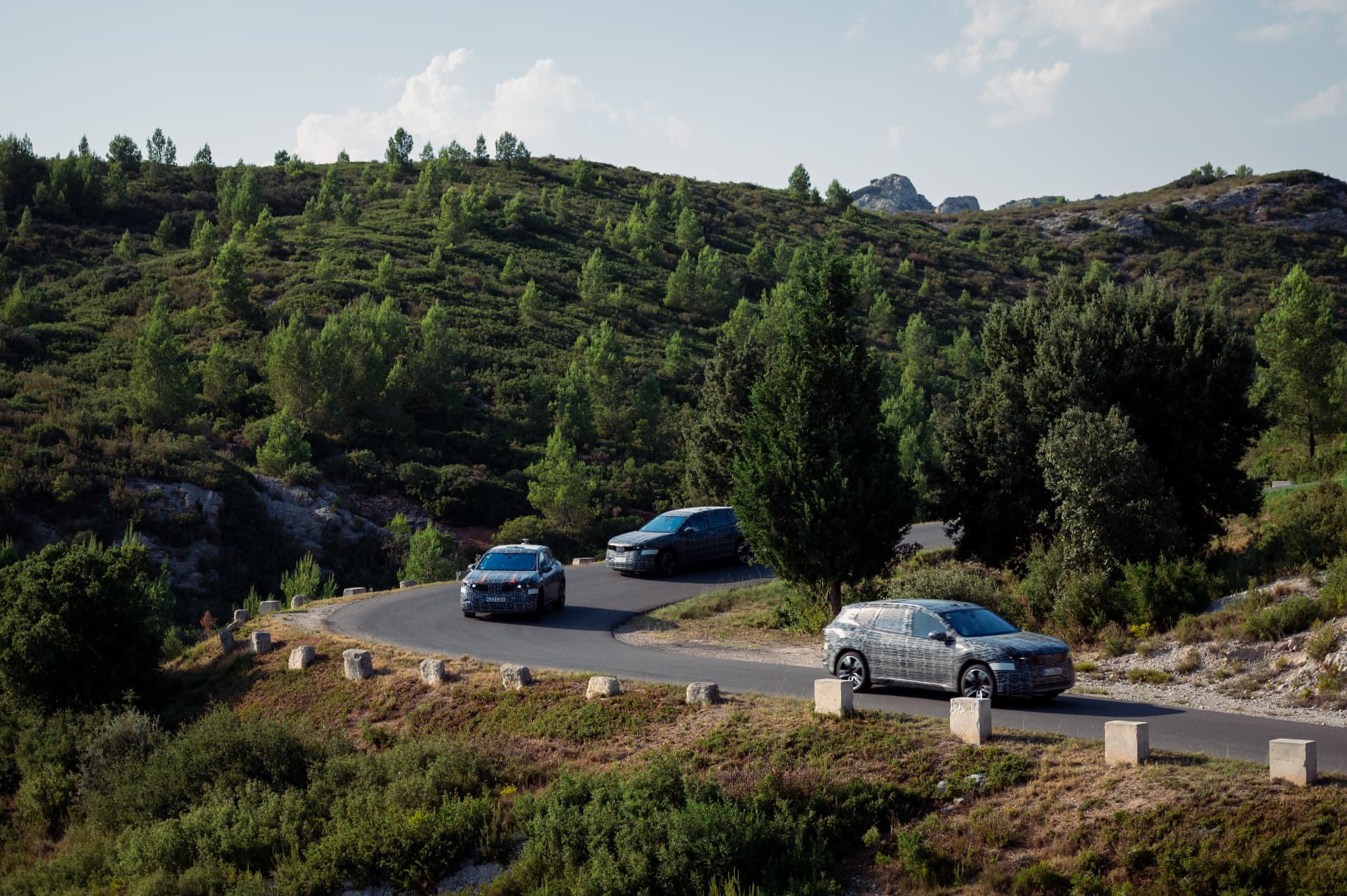 Three camouflaged BMW iX3 prototypes on a mountain curve in Provence during testing near Miramas