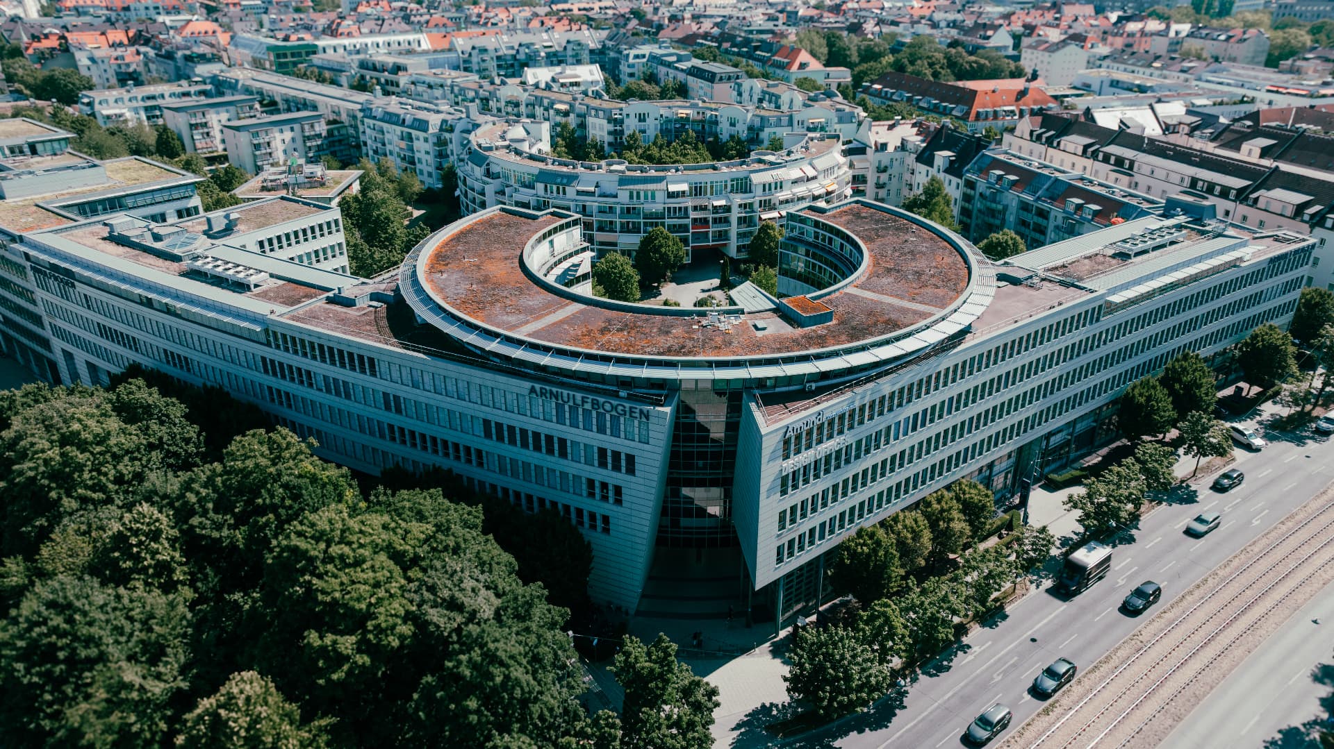 Aerial view of the Arnulfbogen office building in Munich, its curved rooftop ring and inner courtyard framed by trees and the surrounding city