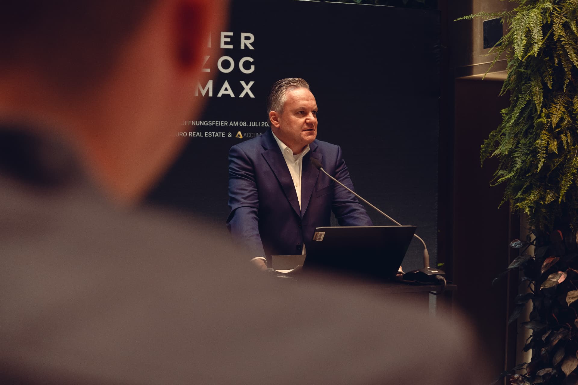 Speaker at the lectern in front of the HERZOG MAX backdrop during the opening ceremony