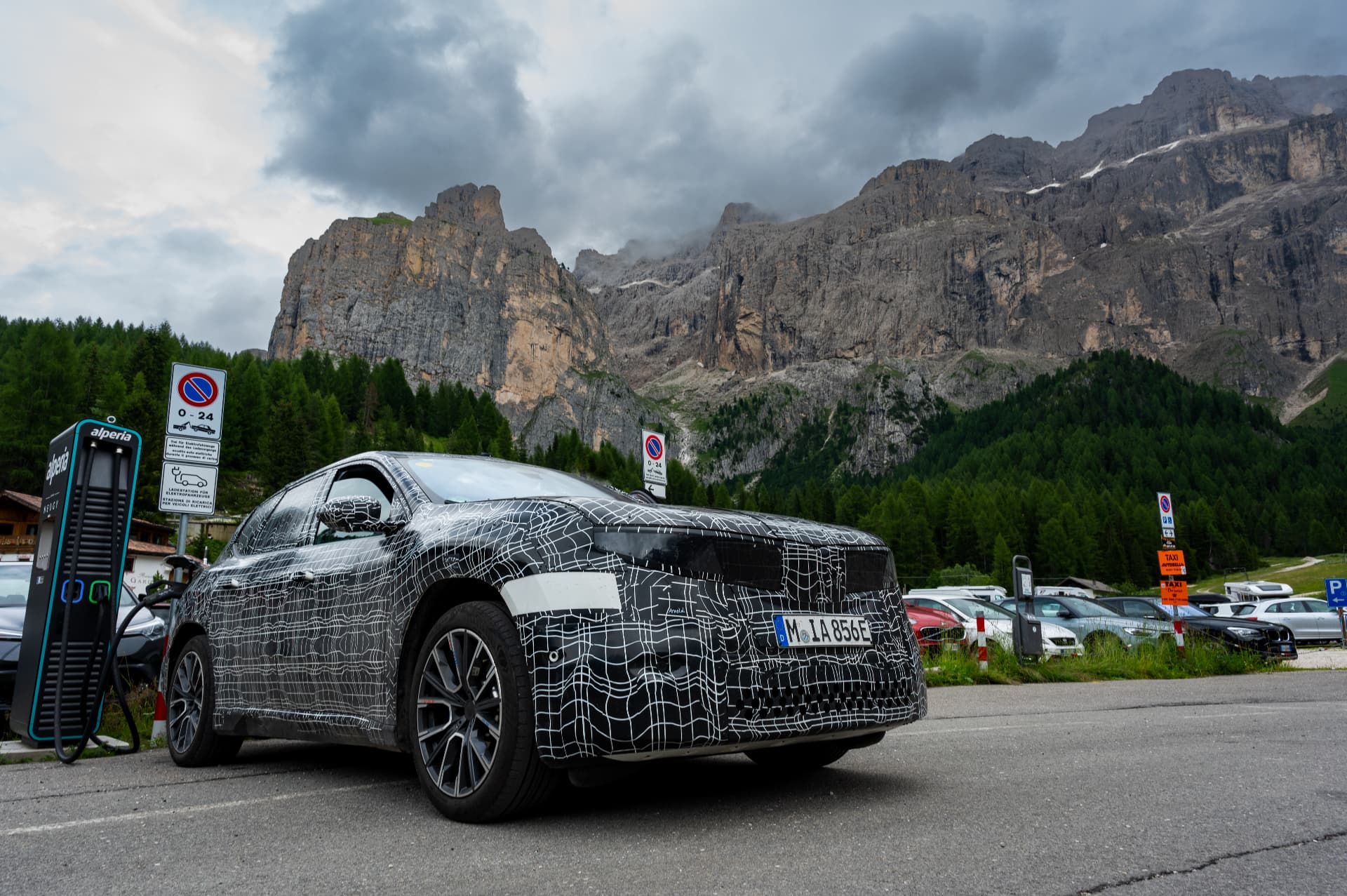 Camouflaged BMW iX3 prototype charging at an electric charging station in front of alpine cliffs