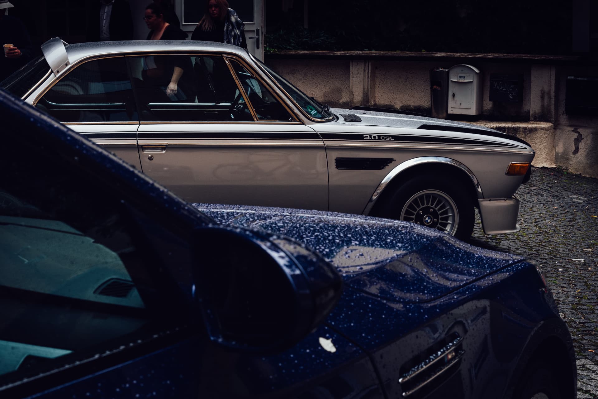 Silver BMW 3.0 CSL in the rain on cobblestones behind a dark blue classic BMW at IAA 2025