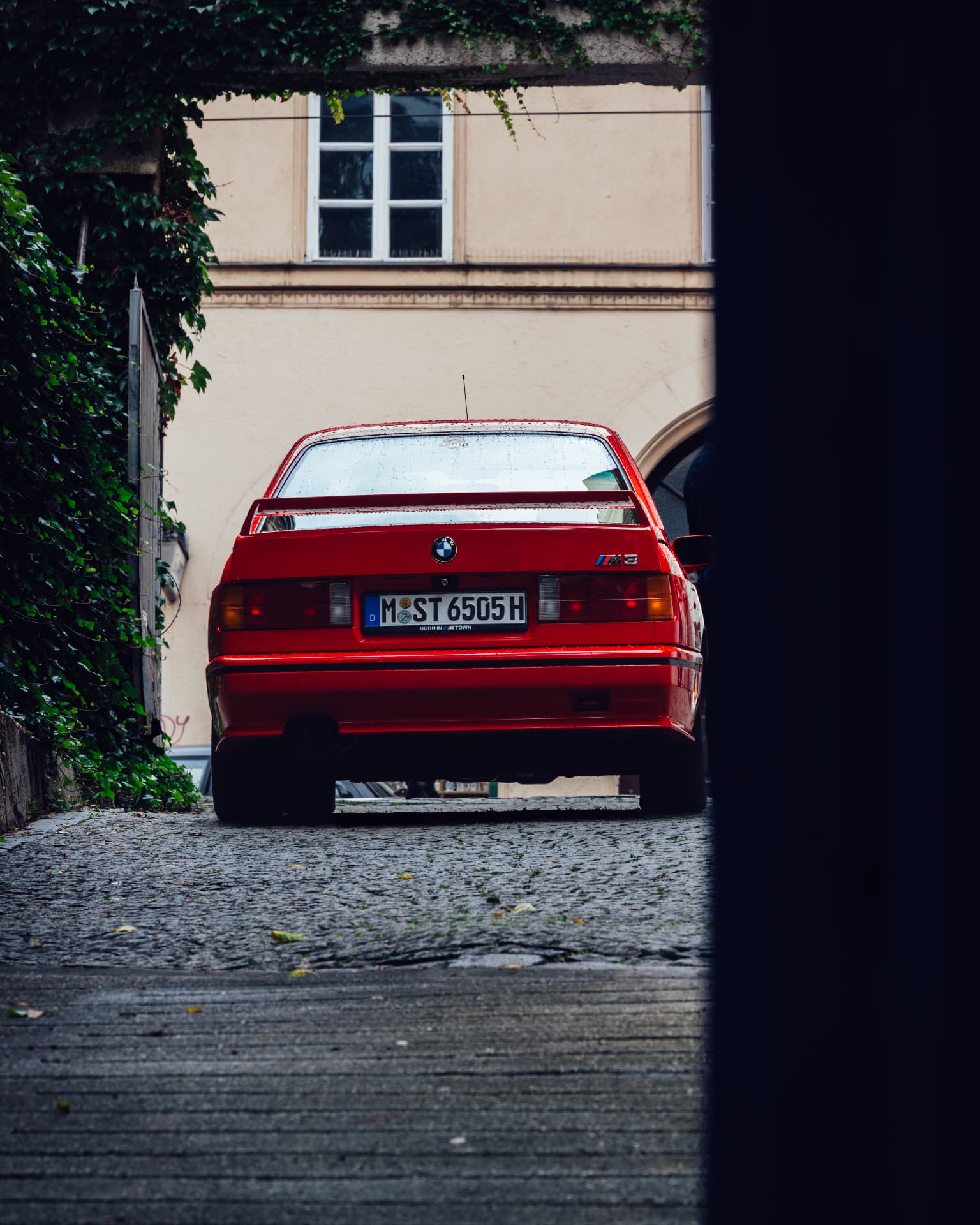 Red BMW M3 E30 parked on a cobblestone driveway framed by ivy and a dark wall