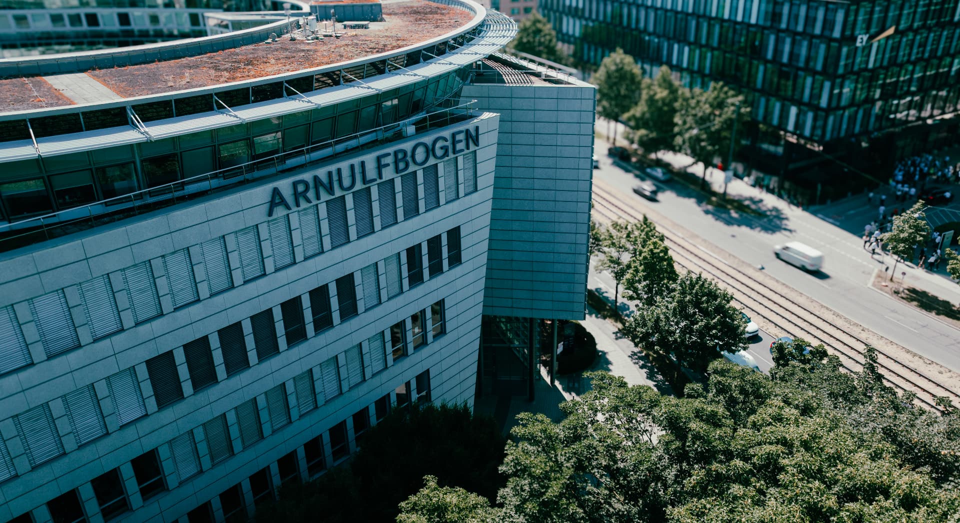 Elevated drone view of the Arnulfbogen lettering on the stone façade, with the street, tram tracks and trees alongside