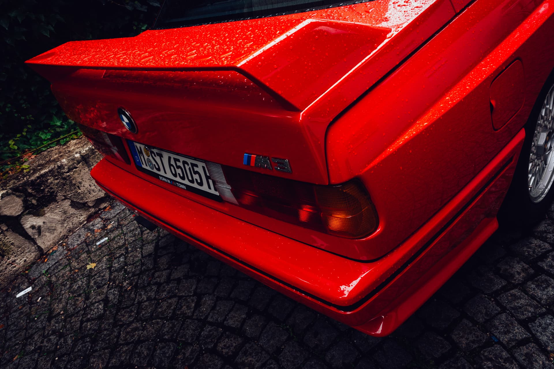 Rear corner of a red BMW M3 E30 with spoiler and M3 badge in the rain