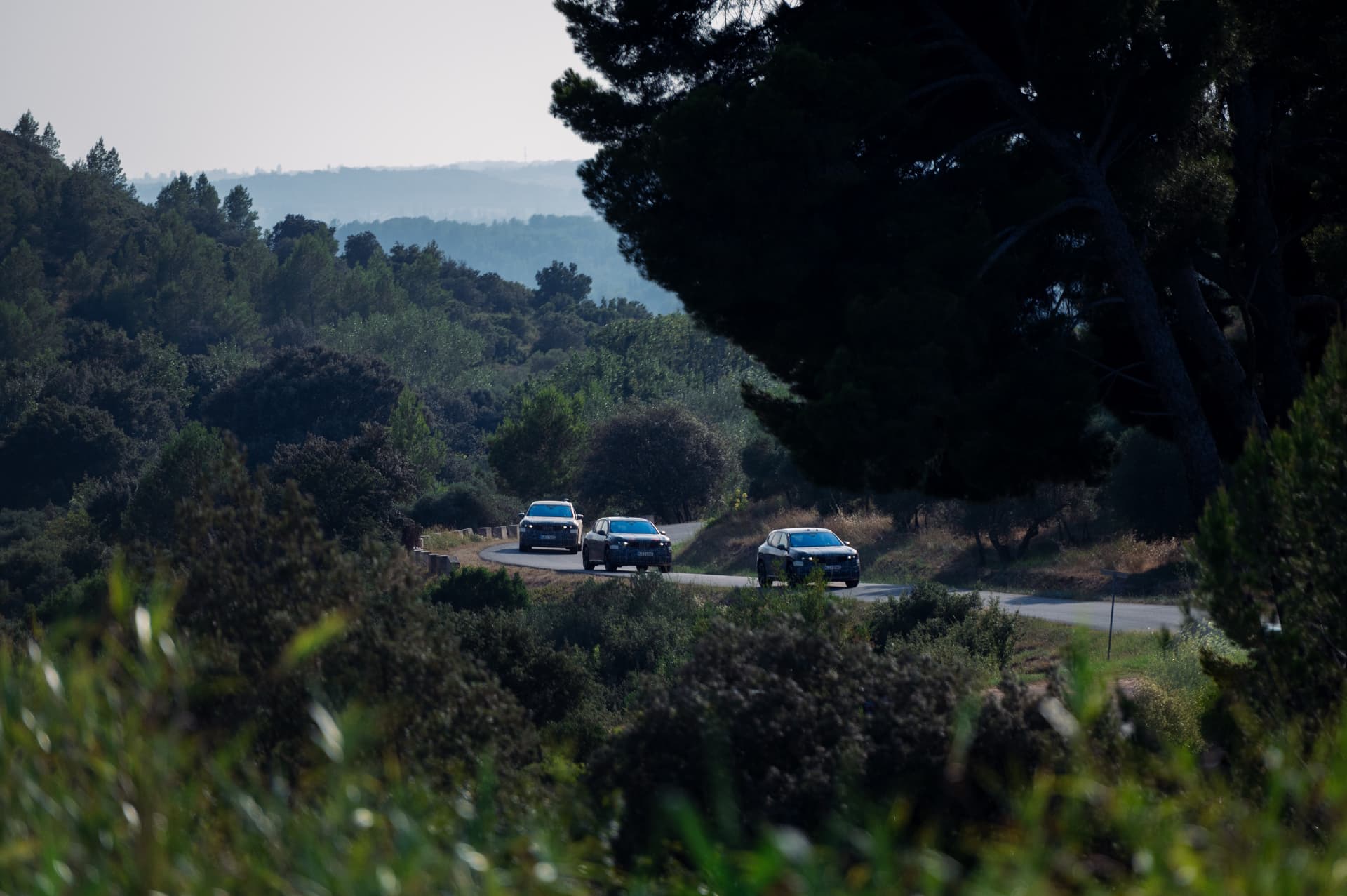 Camouflaged BMW iX3 prototypes approaching on a winding road through wooded hills during testing