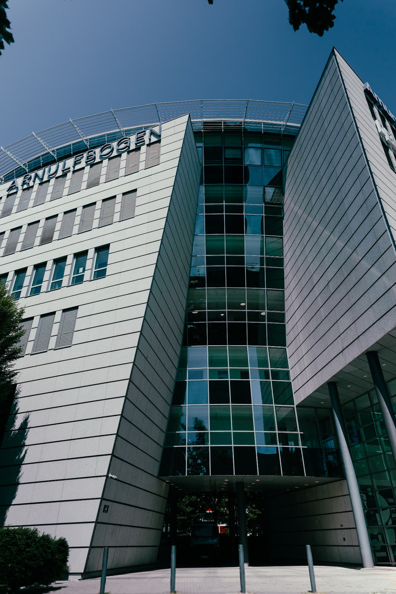 Upward view of the Arnulfbogen entrance, a glazed atrium between two angled stone façades beneath a deep blue sky